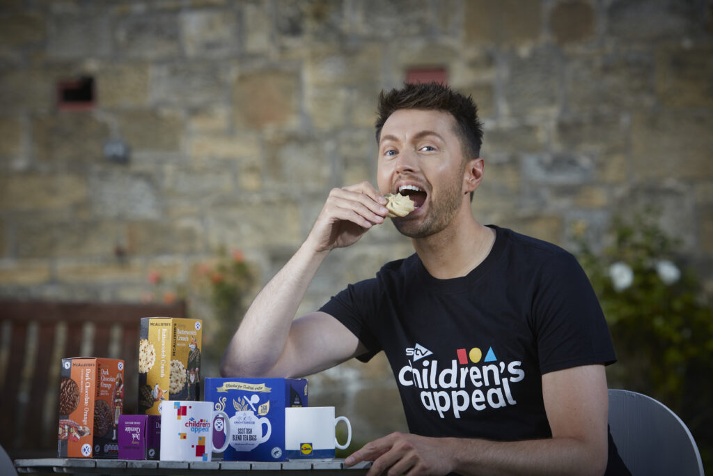A picture of Sean Batty wearing an STV Appeal top eating a biscuit. There are a range of teas and biscuits on the table.
